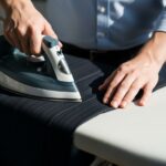 Osoba žehli tmavé nohavice na žehliacej doske v profesionálnom prostredí pod denným svetlom.